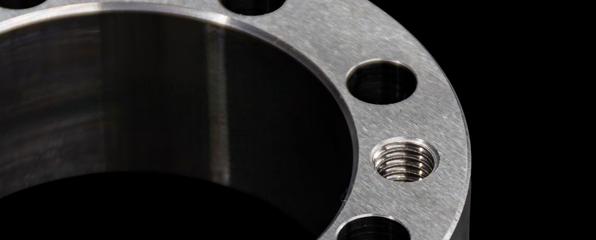 Vergrößerte Ansicht eines geschliffenen Metallrings mit mehreren Bohrungen, darunter eine mit Gewinde. Die Oberfläche ist glatt und präzise bearbeitet, der Hintergrund ist schwarz, wodurch das Metall deutlich hervorsticht.